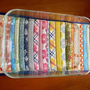 May include: A rectangular glass baking dish rests on a colorful quilted hot pad. The hot pad features vertical strips of patterned fabric in various colors, including blue, pink, and yellow. The dish is empty and sits on a wooden surface.