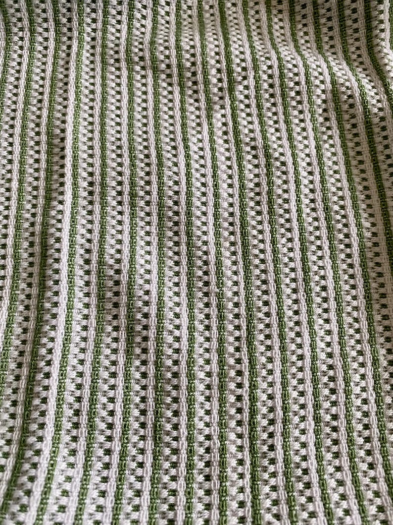 May include: Close-up of a textured fabric with a striped pattern. The fabric features alternating vertical stripes of white and green. The weave creates a grid-like appearance with small rectangular openings throughout the material.