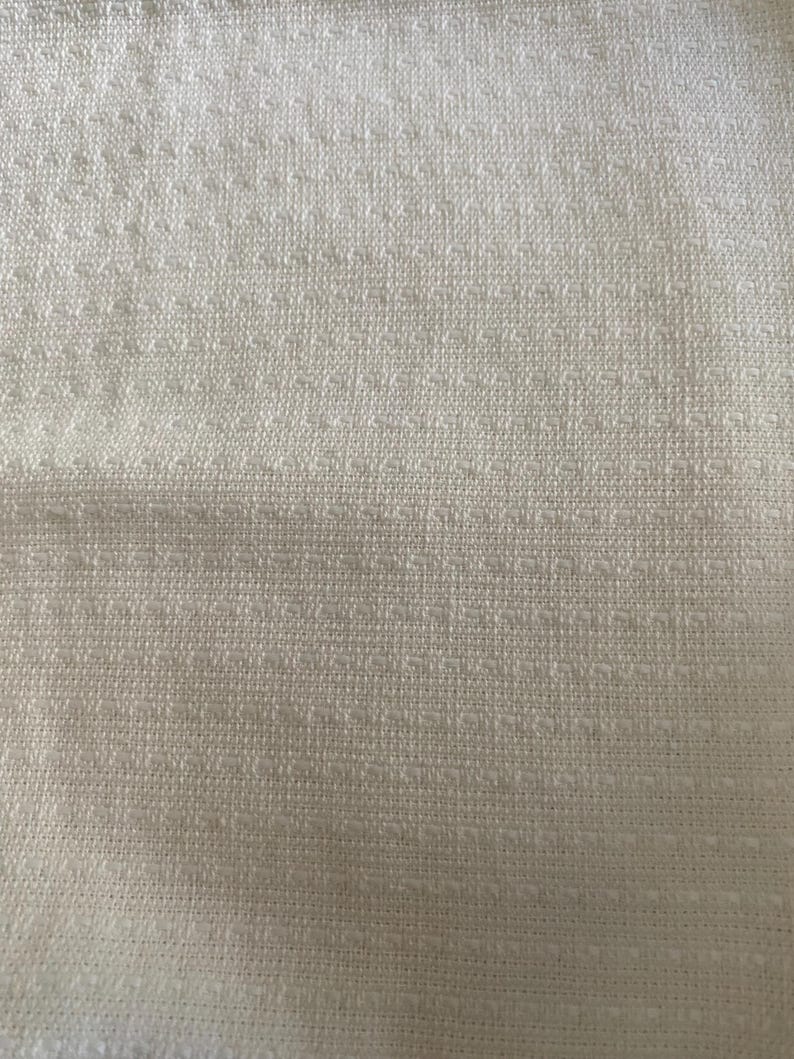 May include: Close-up of a textured, off-white fabric. The material has a woven appearance with a subtle, repeating pattern of small, raised squares. The overall texture is soft and inviting, suggesting a comfortable textile.