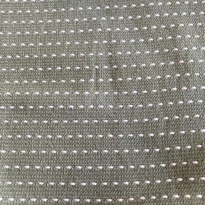 May include: Close-up of a textured fabric in a muted olive green colour. The fabric features a repeating pattern of small, evenly spaced white stitches, creating a subtle, dotted design across the surface.