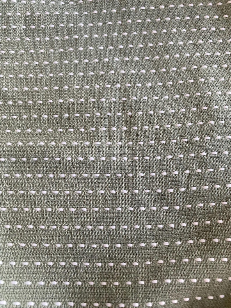 May include: Close-up of a textured fabric in a muted olive green colour. The fabric features a repeating pattern of small, evenly spaced white stitches, creating a subtle, dotted design across the surface.
