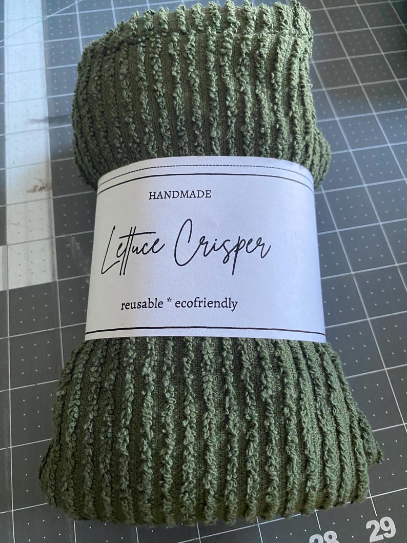 May include: An olive green Lettuce Crisper made of textured fabric, rolled up and ready for use. A white label wrapped around the crisper reads "HANDMADE Lettuce Crisper reusable * ecofriendly" in black script.