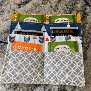May include: A handmade tea bag organizer with multiple pockets. The organizer is made of fabric with various patterns, including a gray geometric design, blue triangles, and yellow floral accents. Tea bags from Twinings, Bigelow, and Choice are visible.