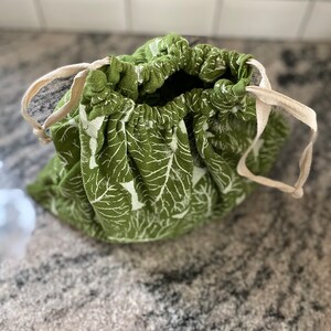 May include: A green fabric drawstring bag featuring a white leaf design. The bag has a natural-coloured drawstring closure. The bag is open, showing the inside.