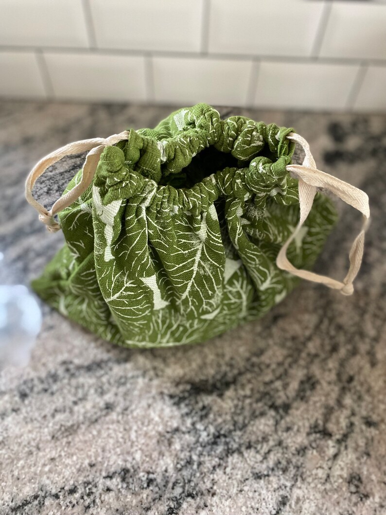 May include: A green fabric drawstring bag featuring a white leaf design. The bag has a natural-coloured drawstring closure. The bag is open, showing the inside.