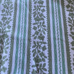 May include: A close-up of a textured fabric featuring a repeating pattern of vertical stripes and floral designs. The stripes alternate between white and a muted green, with the floral elements in a similar green hue. The fabric appears to be woven.