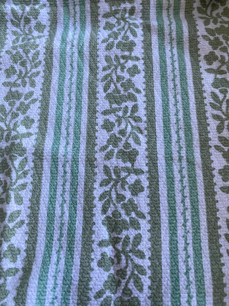 May include: A close-up of a textured fabric featuring a repeating pattern of vertical stripes and floral designs. The stripes alternate between white and a muted green, with the floral elements in a similar green hue. The fabric appears to be woven.