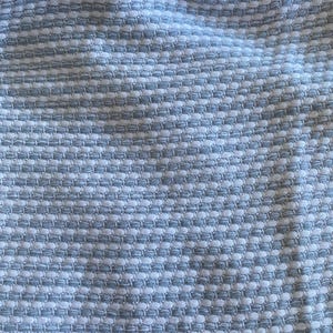 May include: Close-up of a textured fabric featuring a repeating pattern of light blue and white squares. The fabric has a woven appearance, creating a subtle, raised texture. The overall effect is a soft, yet structured design.