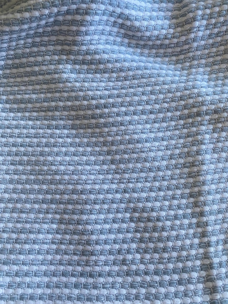 May include: Close-up of a textured fabric featuring a repeating pattern of light blue and white squares. The fabric has a woven appearance, creating a subtle, raised texture. The overall effect is a soft, yet structured design.