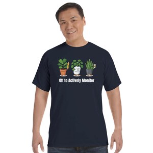 Puede incluir: Camiseta azul marino con un gráfico de tres plantas en macetas, cada una con un accesorio único. El texto "Off to Actively Monitor" está impreso debajo. La camiseta está hecha de un material suave y cómodo.
