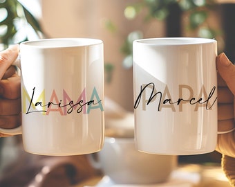 Könnte beinhalten: Zwei weiße Keramikbecher mit Henkeln. Der Becher links trägt das Wort "MAMA" in bunten Buchstaben mit dem Namen "Larissa" darunter. Der Becher rechts trägt das Wort "PAPA" in braunen Buchstaben mit dem Namen "Marcel" darunter.