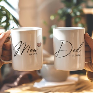 May include: Two white ceramic mugs with black text. The mug on the left says "Mom Est. 2023" with a small pink butterfly. The mug on the right says "Dad Est. 2023".