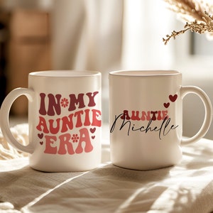 May include: Two white ceramic mugs with pink and red text. One mug says "In My Auntie Era" with a flower and hearts. The other mug says "Auntie Michelle" with a heart.