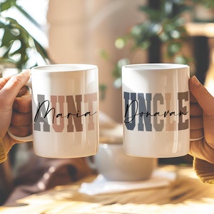May include: Two white ceramic mugs with the words "Aunt Maria" and "Uncle Donovan" printed on them in black and gray lettering. The mugs are held by two hands, with the mugs facing each other.