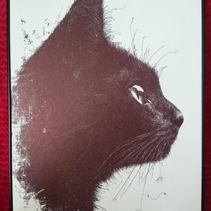 May include: A black and white line art print of a cat's head in profile. The cat's fur is depicted with fine lines, creating a textured effect. The print is framed in a black frame.
