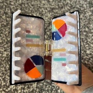 May include: A two-part hair clip with a pearlescent finish and black trim. Each side features colorful pie chart and bar graph designs. The clip is held together by a gold-colored spring mechanism.