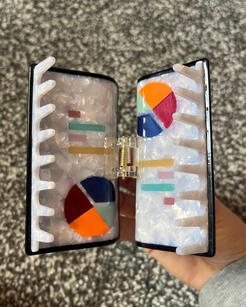 May include: A two-part hair clip with a pearlescent finish and black trim. Each side features colorful pie chart and bar graph designs. The clip is held together by a gold-colored spring mechanism.