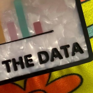 May include: A rectangular, white and black accessory with the text "ME THE DATA" in black. The accessory features a bar graph design with yellow, pink, and green bars.