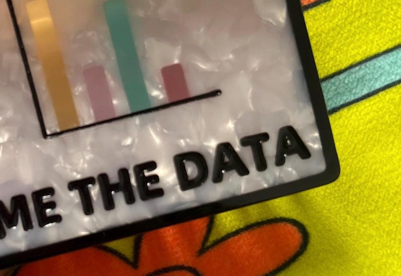 May include: A rectangular, white and black accessory with the text "ME THE DATA" in black. The accessory features a bar graph design with yellow, pink, and green bars.