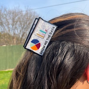 May include: A black and white hair clip with a graphic of a pie chart and bar graph. The text "SHOW ME THE DATA" is printed on the clip. The clip is worn in the hair.