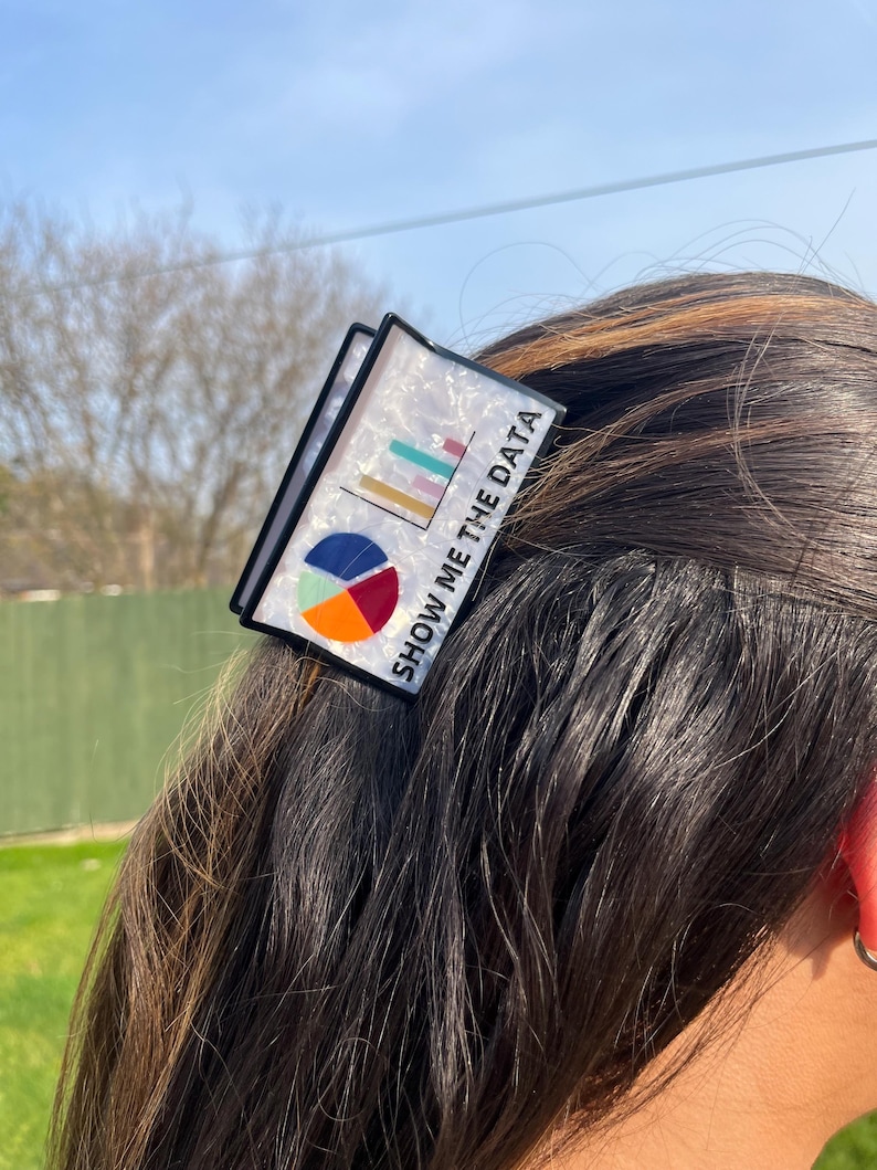 May include: A black and white hair clip with a graphic of a pie chart and bar graph. The text "SHOW ME THE DATA" is printed on the clip. The clip is worn in the hair.