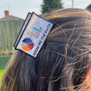 May include: A black and white hair clip with the text "SHOW ME THE DATA". The clip features a colorful bar graph and pie chart design. The clip is set against a background of brown hair.