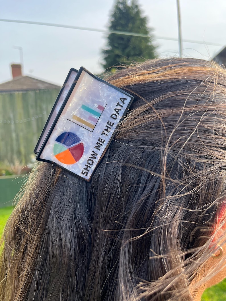 May include: A black and white hair clip with the text "SHOW ME THE DATA". The clip features a colorful bar graph and pie chart design. The clip is set against a background of brown hair.