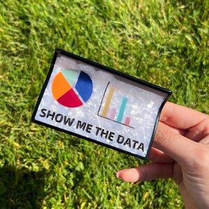 May include: A rectangular, black-framed pin with a white background featuring a colorful pie chart and bar graph. The text "SHOW ME THE DATA" is printed in black. The pin is held against a background of green grass.
