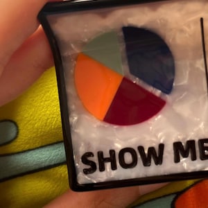 May include: A rectangular, black-framed object with a pie chart design in orange, green, navy, and burgundy. The text "SHOW ME" is printed in bold, black letters at the bottom of the object.