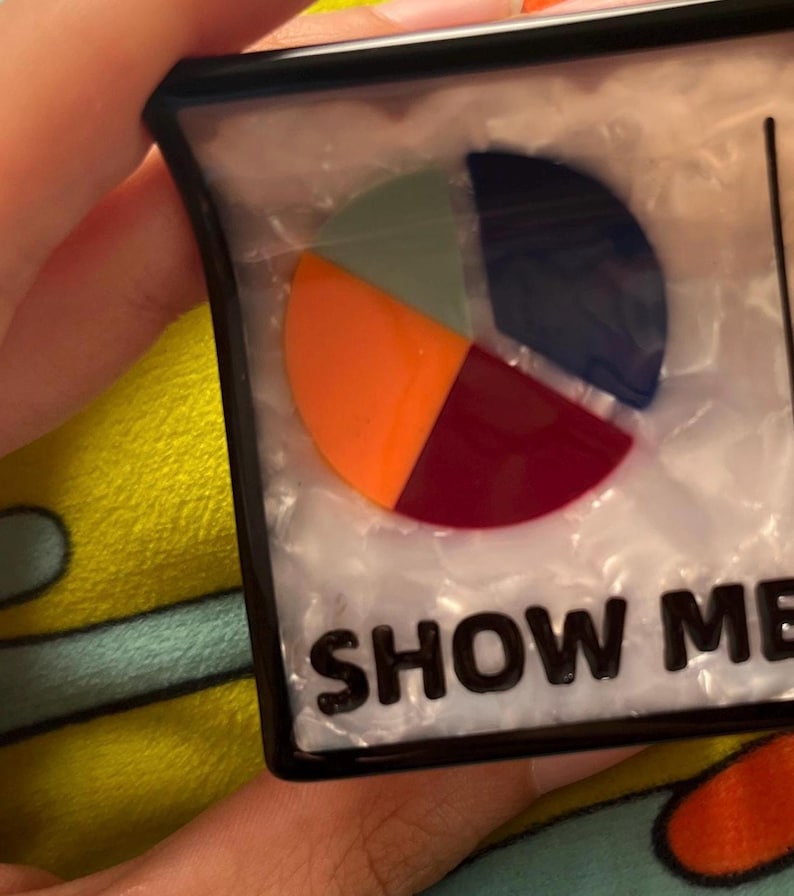 May include: A rectangular, black-framed object with a pie chart design in orange, green, navy, and burgundy. The text "SHOW ME" is printed in bold, black letters at the bottom of the object.