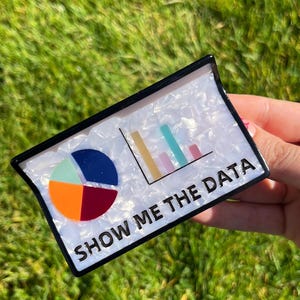 May include: A rectangular pin with a black border and a white, marbled background. The pin features a pie chart and a bar graph, with the text "SHOW ME THE DATA" in bold, white letters.