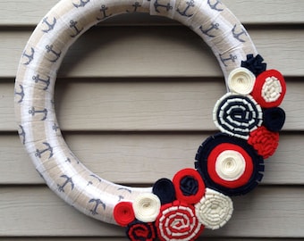 May include: A circular wreath wrapped in beige fabric featuring a repeating dark blue anchor pattern. A matching fabric ribbon hangs from the top. The lower right side is decorated with a cluster of rolled felt flowers in red, white, and navy blue.