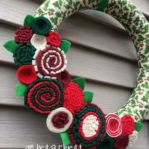 May include: A Christmas wreath with a green and white holly pattern. The wreath is decorated with felt flowers in shades of red, green, white, and burgundy.
