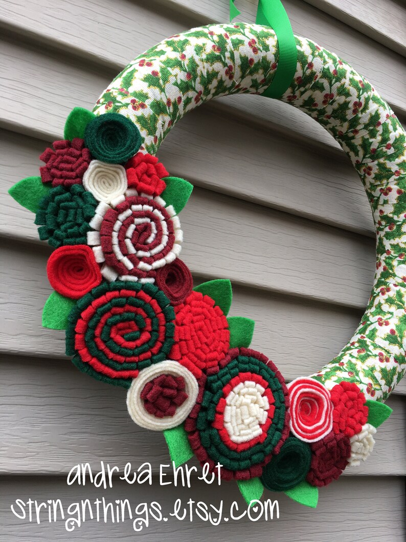 May include: A Christmas wreath with a green and white holly pattern. The wreath is decorated with felt flowers in shades of red, green, white, and burgundy.