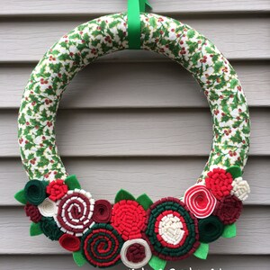 May include: A Christmas wreath made of green and white fabric with a holly pattern. The wreath is decorated with red, white and green felt flowers. The wreath is hanging on a grey wall with a green ribbon.