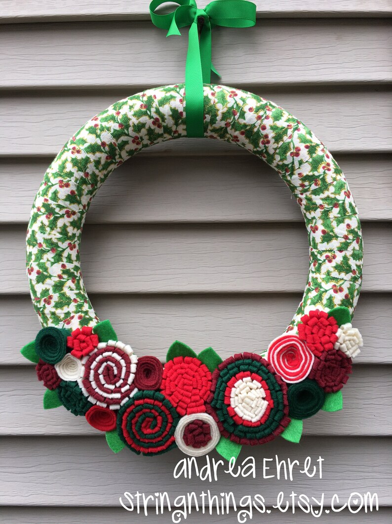 May include: A Christmas wreath made of green and white fabric with a holly pattern. The wreath is decorated with red, white and green felt flowers. The wreath is hanging on a grey wall with a green ribbon.
