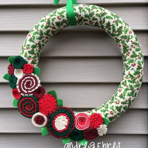 May include: A green felt Christmas wreath with a holly pattern. The wreath is decorated with red, white and green felt flowers. The wreath is hanging on a grey wall. The text "andrea Ehret stringthings.etsy.com" is printed below the wreath.