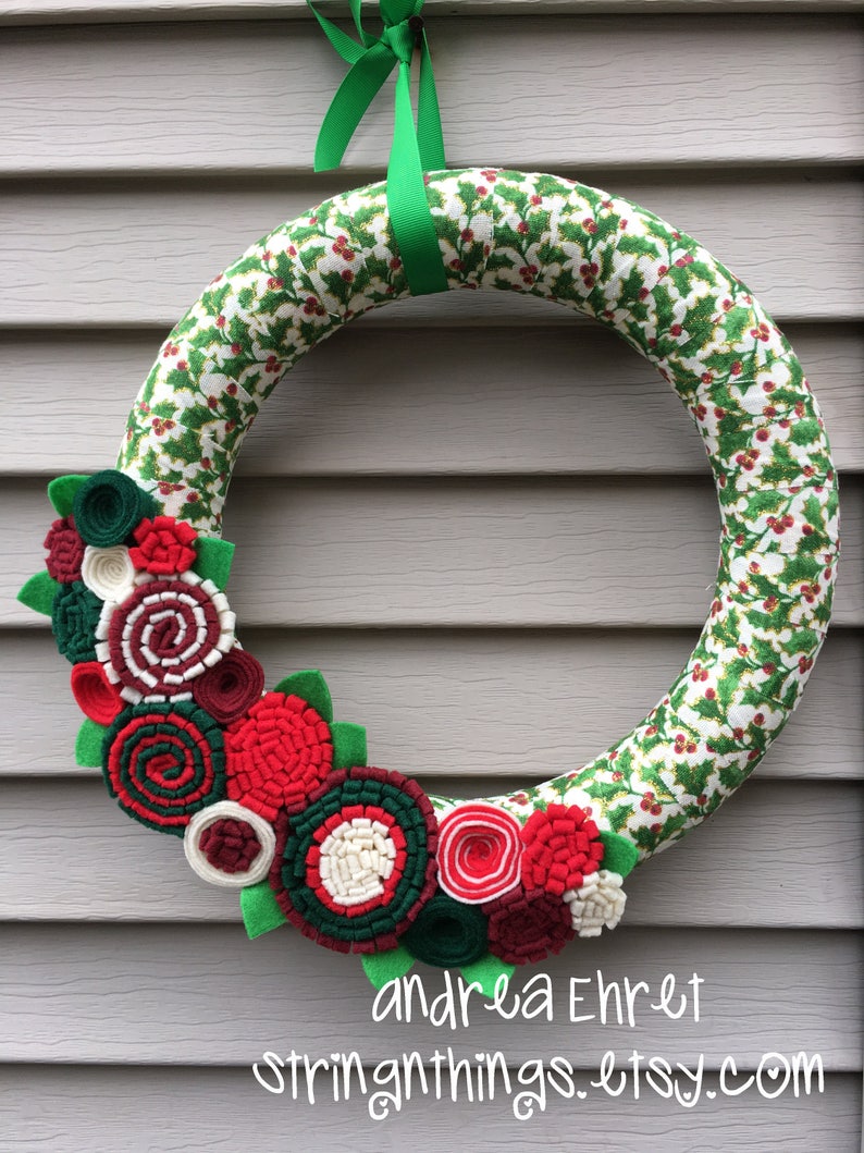 May include: A green felt Christmas wreath with a holly pattern. The wreath is decorated with red, white and green felt flowers. The wreath is hanging on a grey wall. The text "andrea Ehret stringthings.etsy.com" is printed below the wreath.