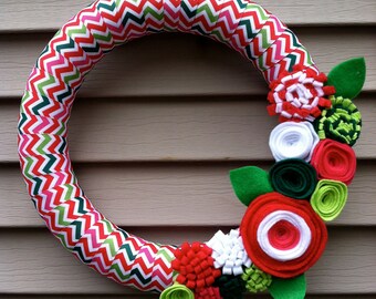 May include: A handmade wreath with a chevron pattern in red, green, and white. The wreath is decorated with felt flowers in red, white, green, and lime green, and green felt leaves. This festive wreath is perfect for seasonal decor.
