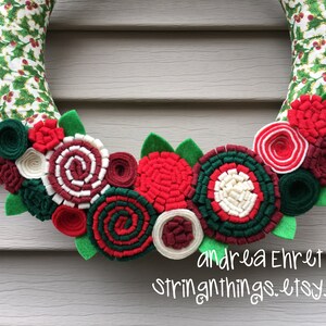 May include: A Christmas wreath made of green and white fabric with red and white felt flowers and green felt leaves. The wreath is decorated with red, white, and green felt rosettes. The text "andrea Ehret stringthings.etsy.com" is visible at the bottom of the image.