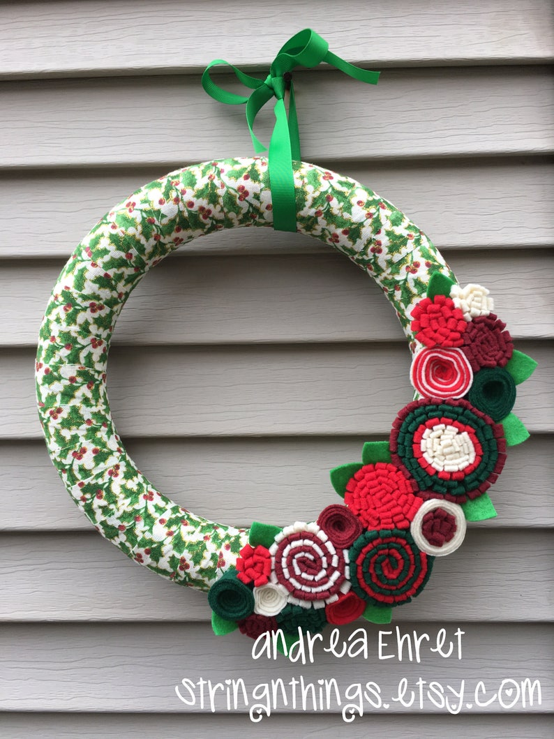 May include: A Christmas wreath made of green and white fabric with a holly pattern. The wreath is decorated with red, white and green felt flowers. The wreath is tied with a green ribbon and has the text "andrea Ehret stringthings.etsy.com" at the bottom.
