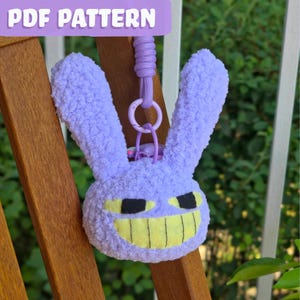 Puede incluir: Un llavero de cabeza de conejo de peluche difuso de color morado claro con orejas largas y una amplia sonrisa amarilla con dientes negros cosidos y ojos negros rectangulares. El peluche está unido a un lazo de cordón morado y un anillo de plástico.
