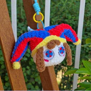 May include: A plush keychain featuring a white round character with brown hair and blue and red circular eyes. The character wears a jester hat in red, blue, and yellow with yellow pom-poms at the ends, attached to a blue cord and yellow ring.