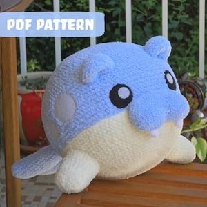 May include: A soft toy resembling a cartoon character, predominantly light blue and cream. It has large black eyes, white teeth, and a rounded form. The text "PDF PATTERN" is displayed on a light blue banner.