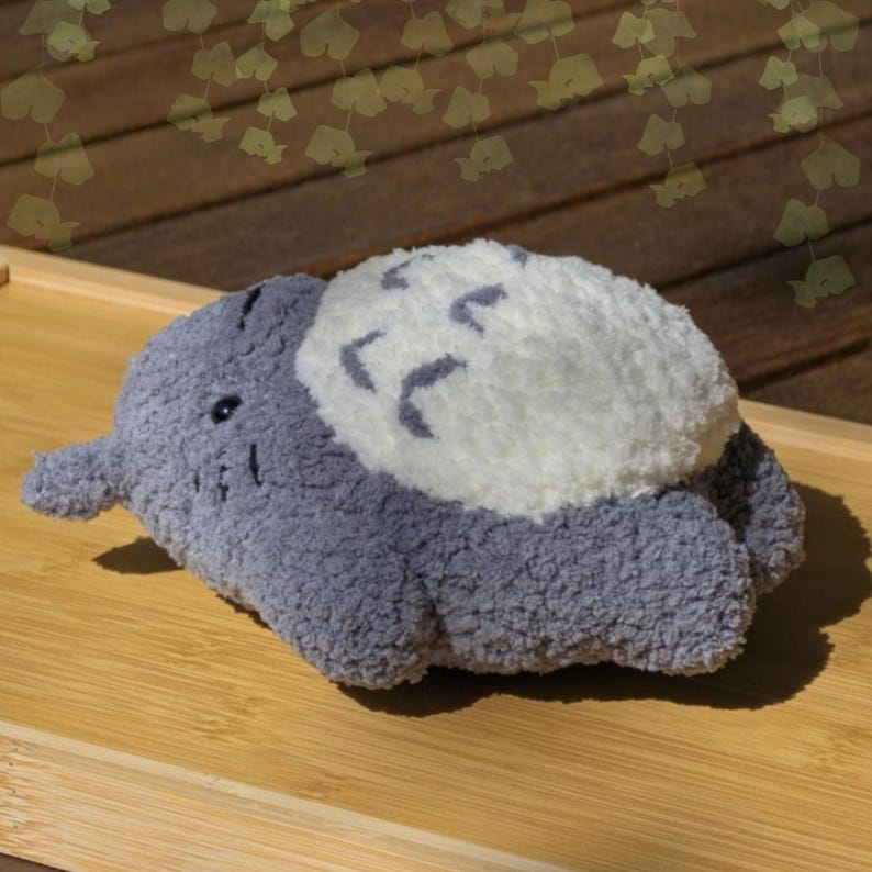 May include: A plush, gray stuffed animal with a white, cloud-like design on its back. The toy has small black eyes and a short trunk. The toy is lying on a light brown wooden surface.