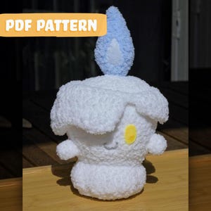 May include: A plush, white, candle-shaped toy with a blue flame and a yellow accent. The toy has a smiling face and small arms. The text "PDF PATTERN" is displayed in a yellow banner.