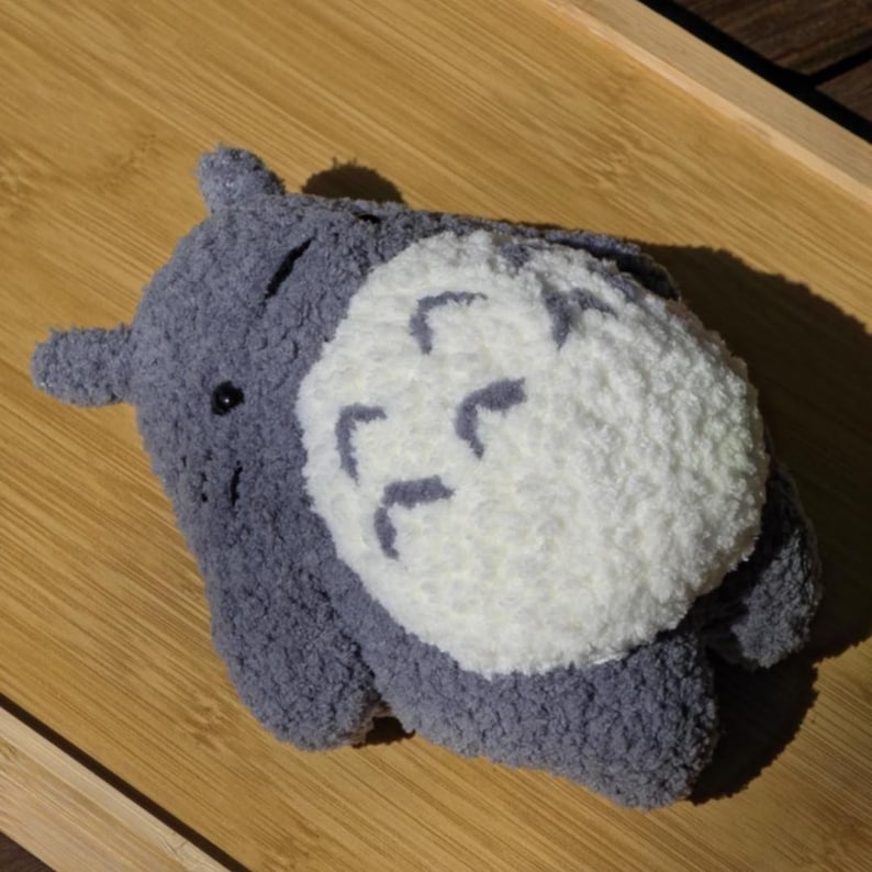 May include: A plush, gray Totoro-inspired character with a white belly and dark gray markings. The soft toy has small ears and a smiling face. The plush is lying on a wooden surface.