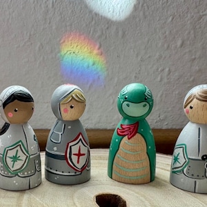May include: Four wooden peg dolls: two knights, a dragon, and a lady. The knights wear gray armor with shields. The dragon is green with a red scarf. The lady wears a white dress. The dolls are hand-painted.