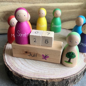 Può includere: Un calendario in legno con figure di legno colorate che rappresentano i giorni della settimana. Il calendario è impostato sul 28 luglio. Le figure sono dipinte in diversi colori, tra cui rosa, giallo, verde, blu e viola. Ogni figura ha un viso dipinto e una parola che rappresenta il giorno della settimana.