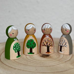 May include: Four wooden peg dolls, each representing a season. The dolls are painted with green, yellow, brown, and gray clothing. Each doll features a tree design depicting spring, summer, autumn, and winter. The dolls are displayed on a wooden surface.
